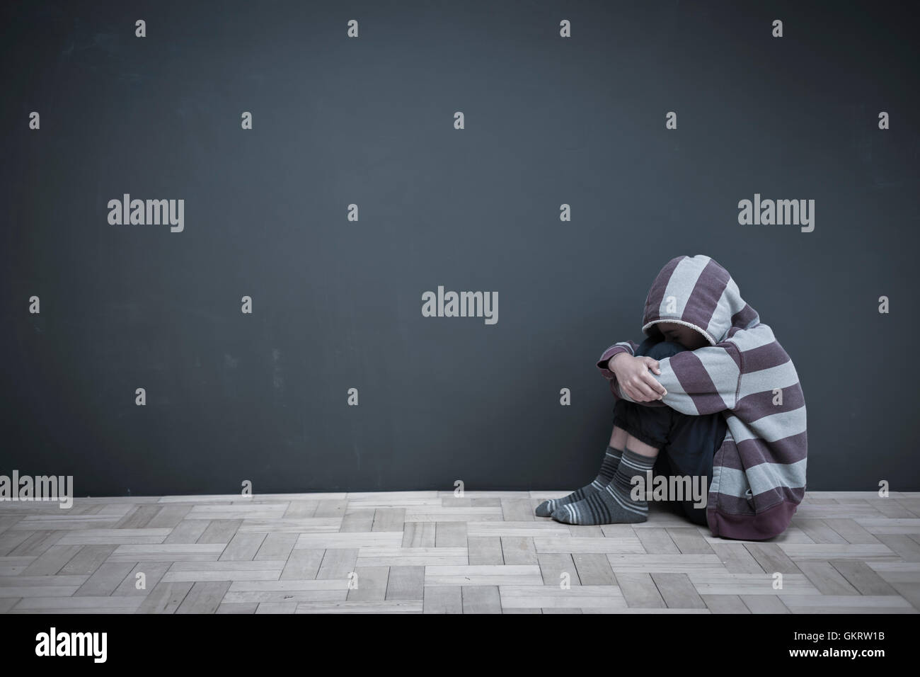 Boy sitting on floor hugging knees with head down Stock Photo