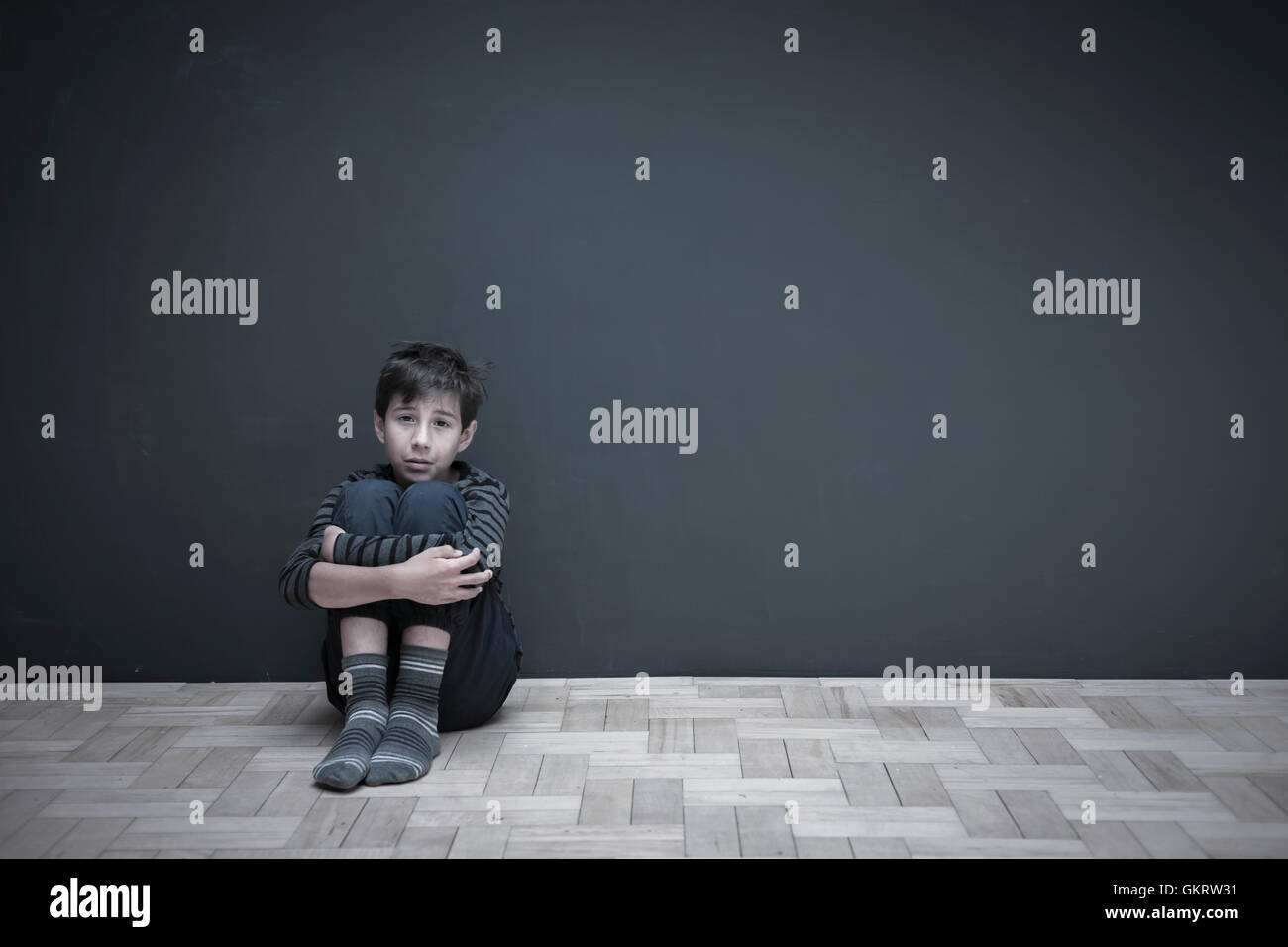 Boy sitting on floor against grey wall hugging knees Stock Photo