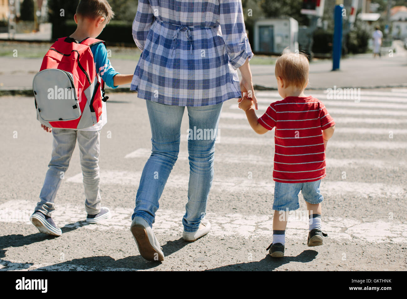 Mother and her children crossing road on way to school. Mother holding her sons hands walking across zebra crossing. Stock Photo