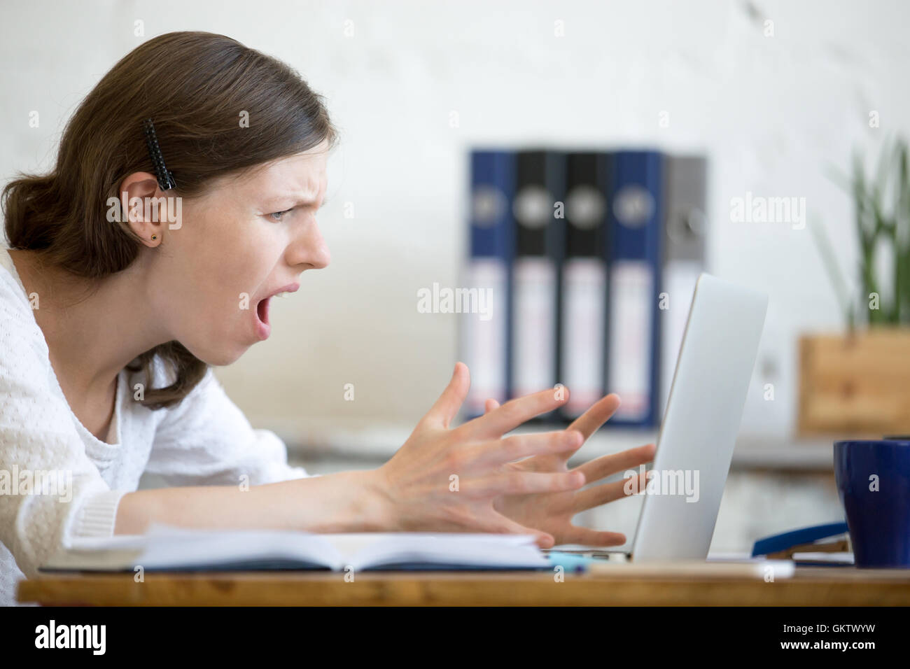 Young stressed businesswoman sitting with laptop and screaming with angry expression. Furious business person feeling irritated Stock Photo