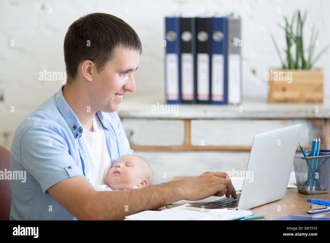 Side view portrait of happy casual young business dad holding newborn cute babe while working on laptop in home office interior. Stock Photo