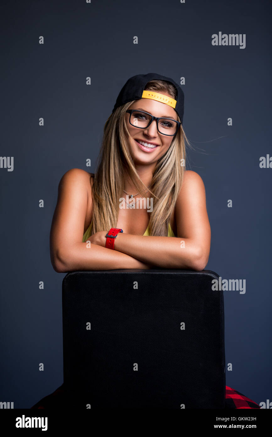 Beautiful girl in cap and glasses sitting on chair, grey backdrop Stock Photo