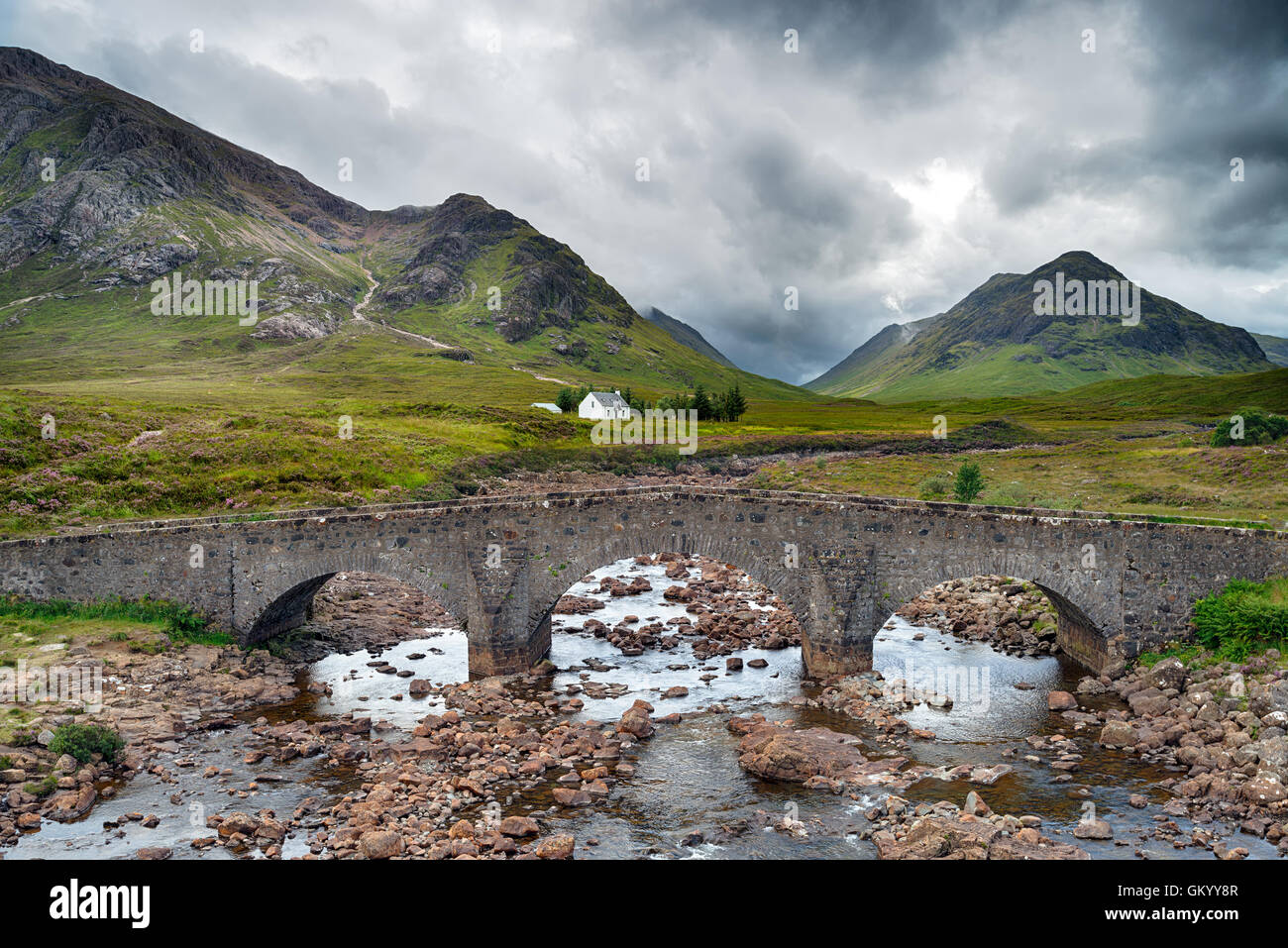 An ancient stone bridge backed by rugged mountains and a lonely cottage under a dramatic moody sky Stock Photo