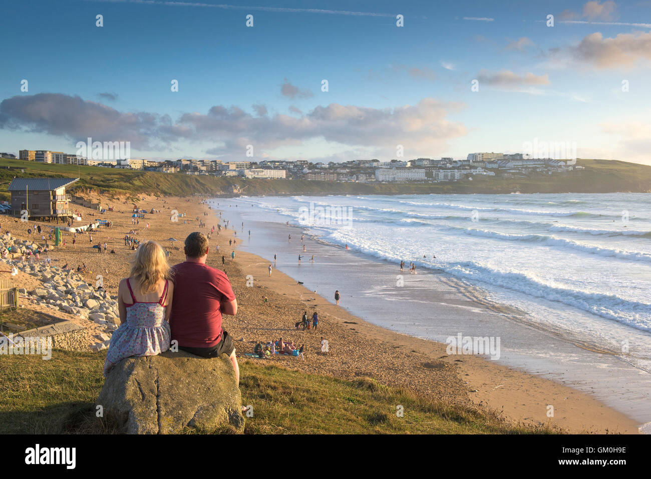 Two holidaymakers sit on a rock and enjoy the sunset over Fistral Beach in Newquay, Cornwall. Stock Photo