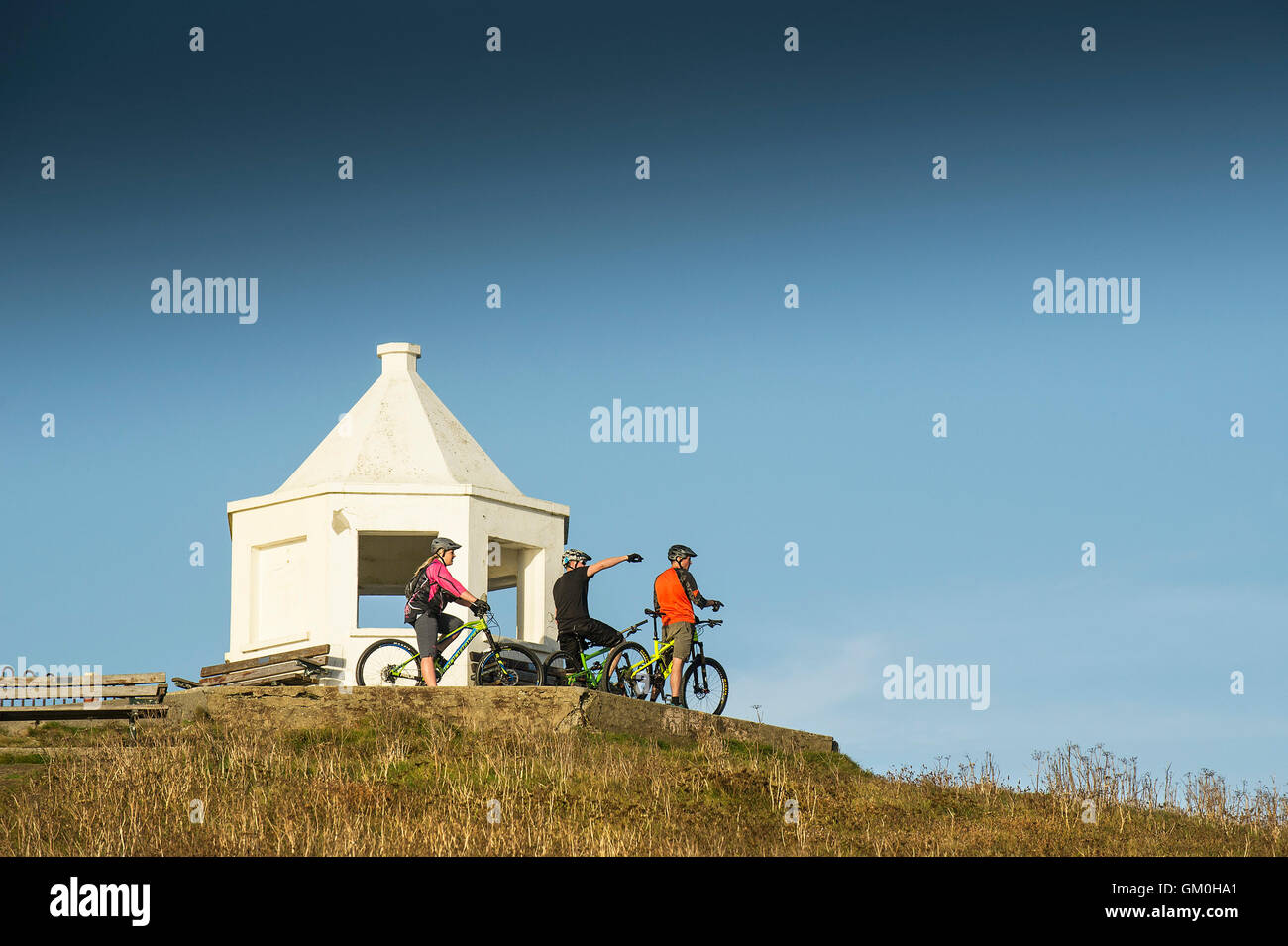 Three mountain bikers stand next to the white lookout building on toip of Towan Headland in Newquay, Cornwall. Stock Photo