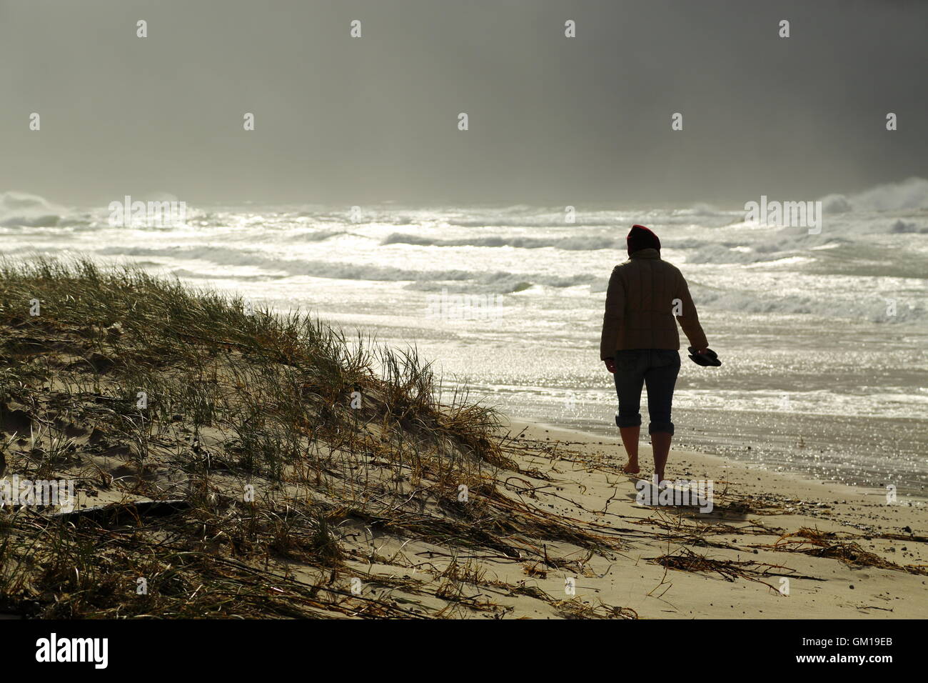 A young lady in her thirties braces against the winter storm as she walks barefoot on a beach at Iluka in NSW, Australia. Stock Photo