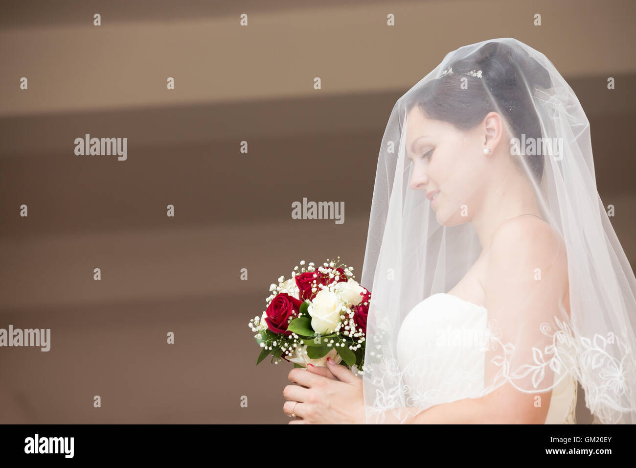 Side view portrait of young beautiful bride posing with her eyes down, holding bridal bouquet with white and red roses Stock Photo