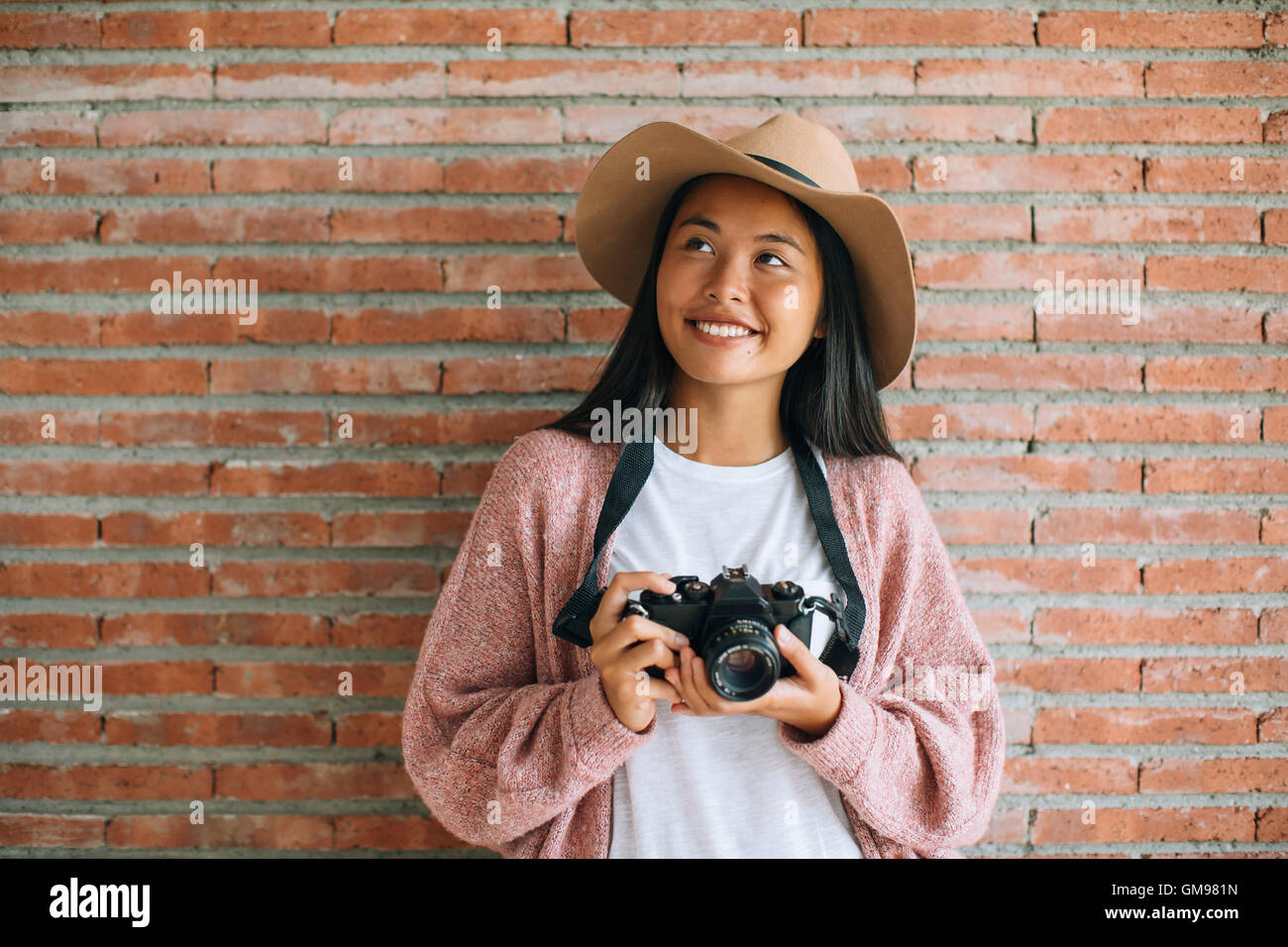 Portrait of smiling young woman with camera in front of brick wall ...