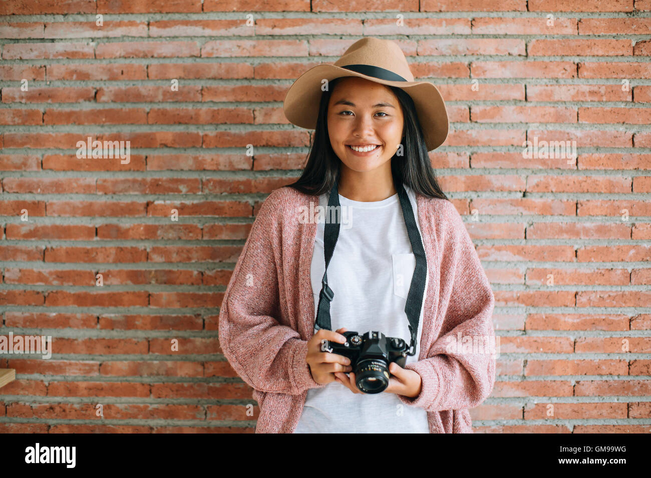 Portrait of smiling young woman with camera in front of brick wall ...