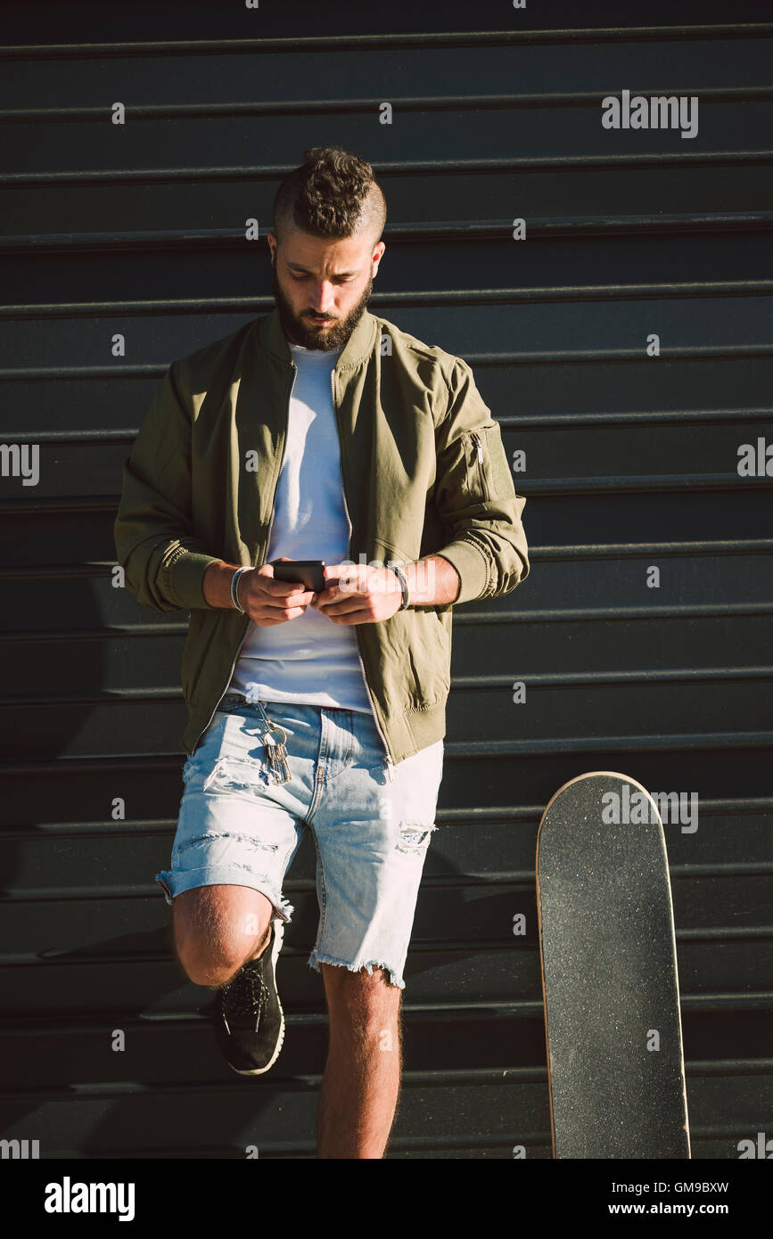 Man with skateboard standing in front of facade looking at cell phone Stock Photo
