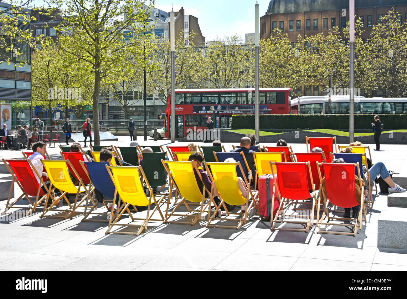 People using colourful deck chairs placed outside office buildings in Central London England UK on a sunny late spring day traffic on busy road beyond Stock Photo