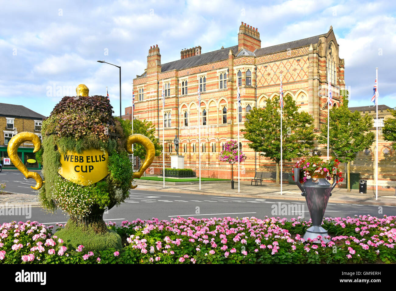Poor replica of Webb Ellis Rugby Cup in flower display opposite statue of William Webb Ellis outside Rugby School buildings Warwickshire England UK Stock Photo