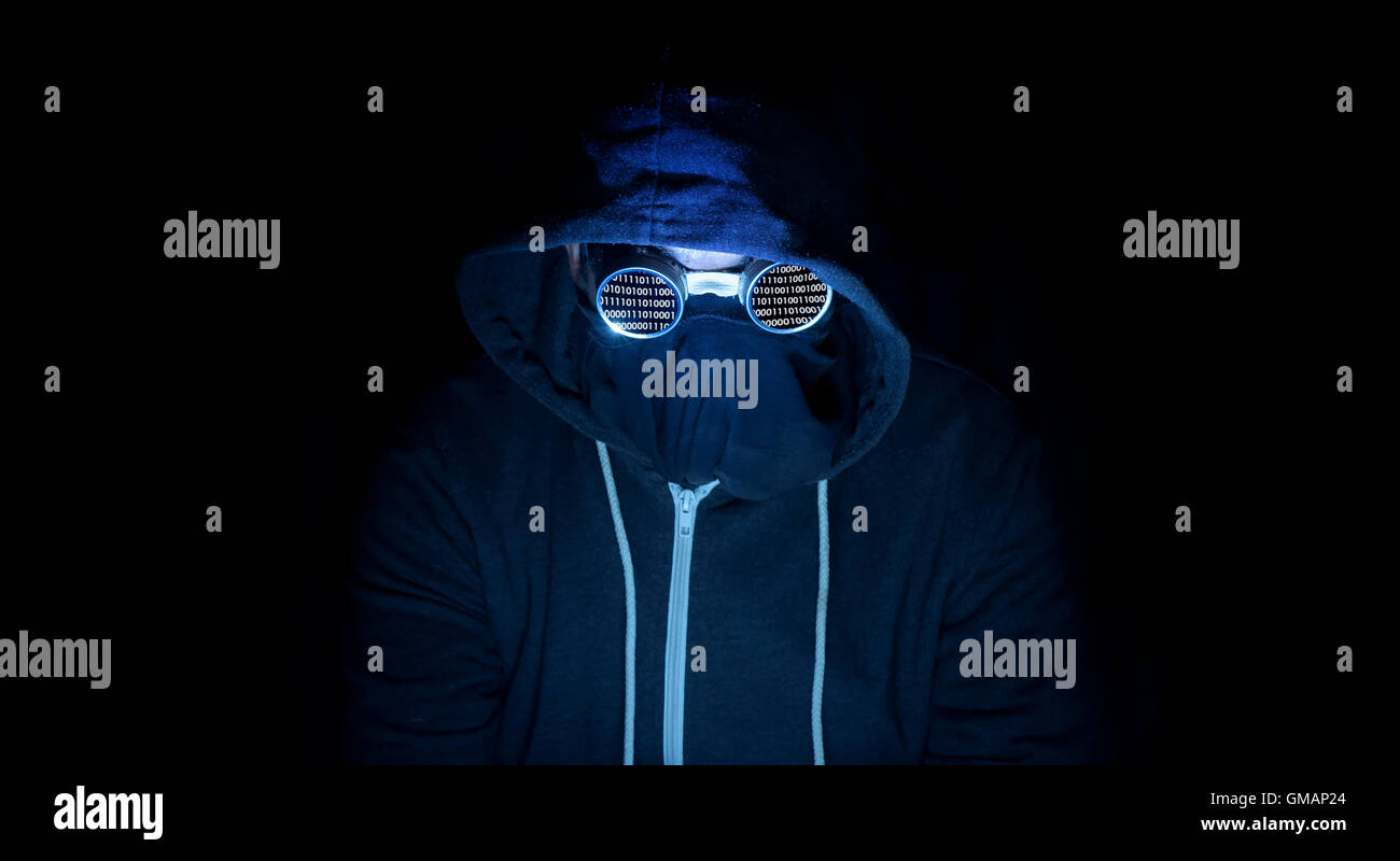 Hooded computer criminal hacker sits in a dark room wearing goggles to conceal his identity as he commits crimes on the internet Stock Photo