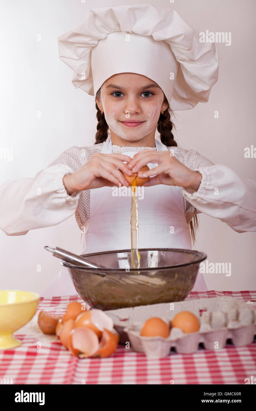 Little cook girl in a white apron breaks eggs in a deep dish, isolated on white background Stock Photo