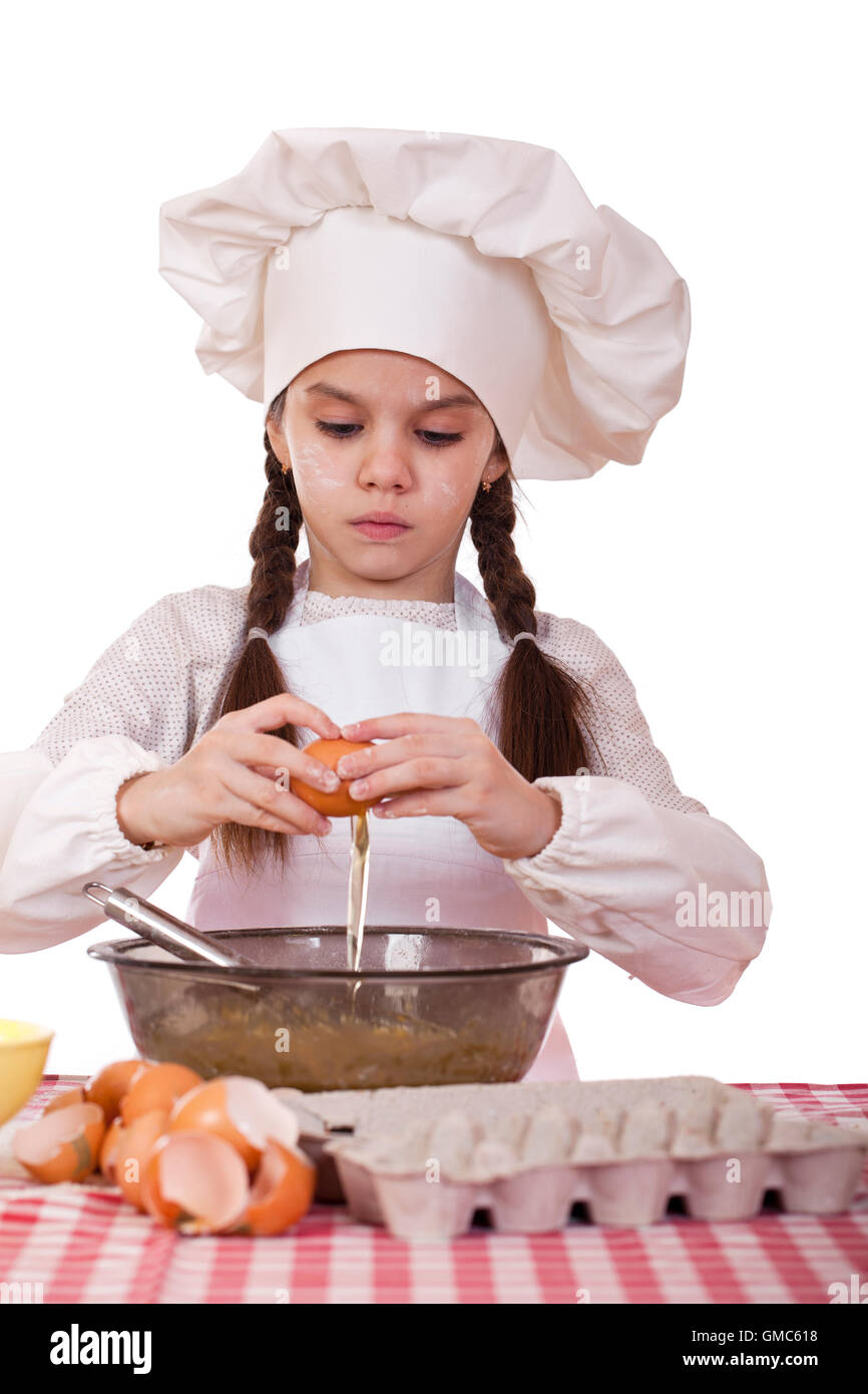 Little cook girl in a white apron breaks eggs in a deep dish, isolated on white background Stock Photo