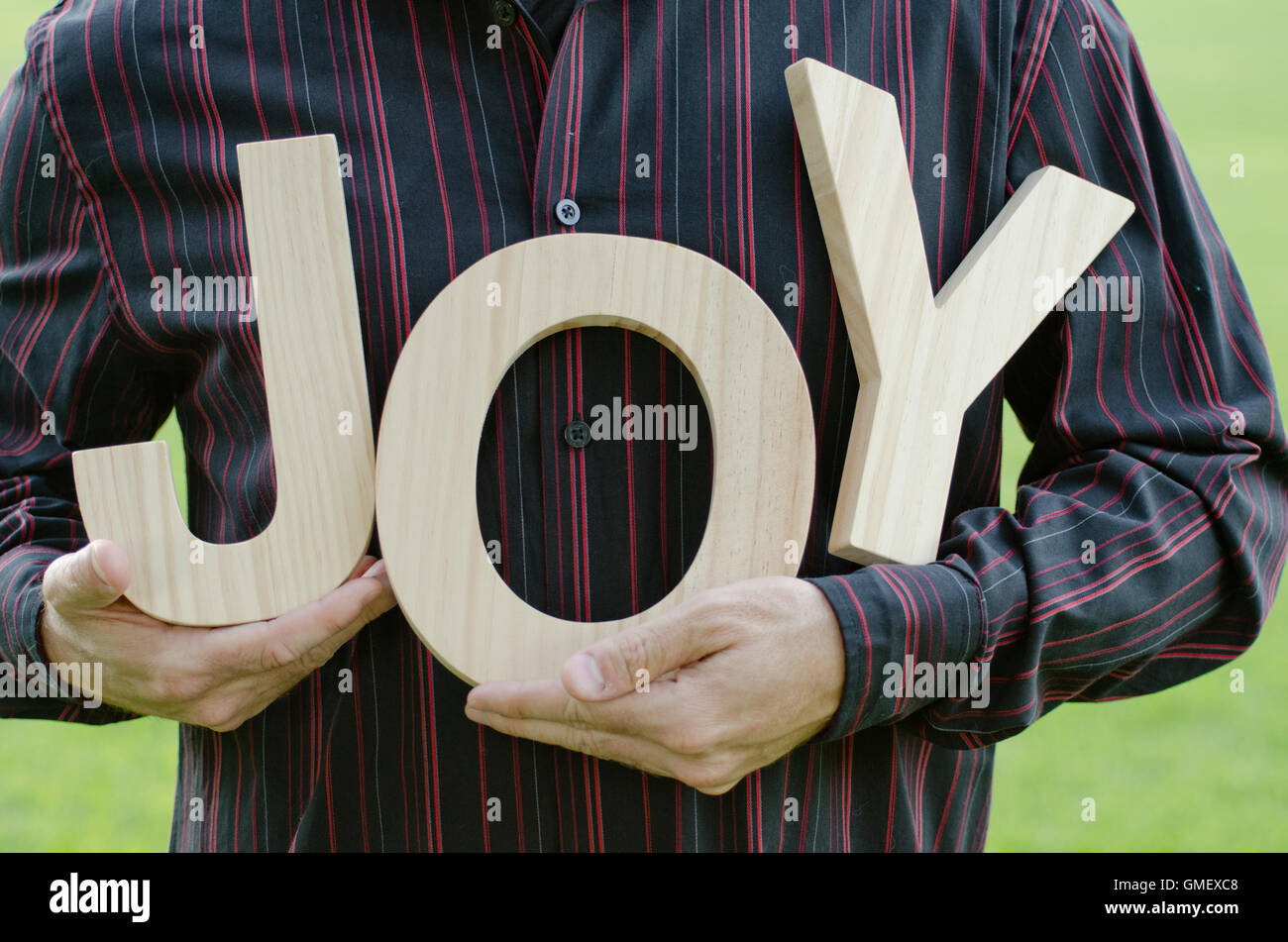 Close up of man's hands holding the wooden letters of the word joy, while wearing a red striped button up shirt. Stock Photo