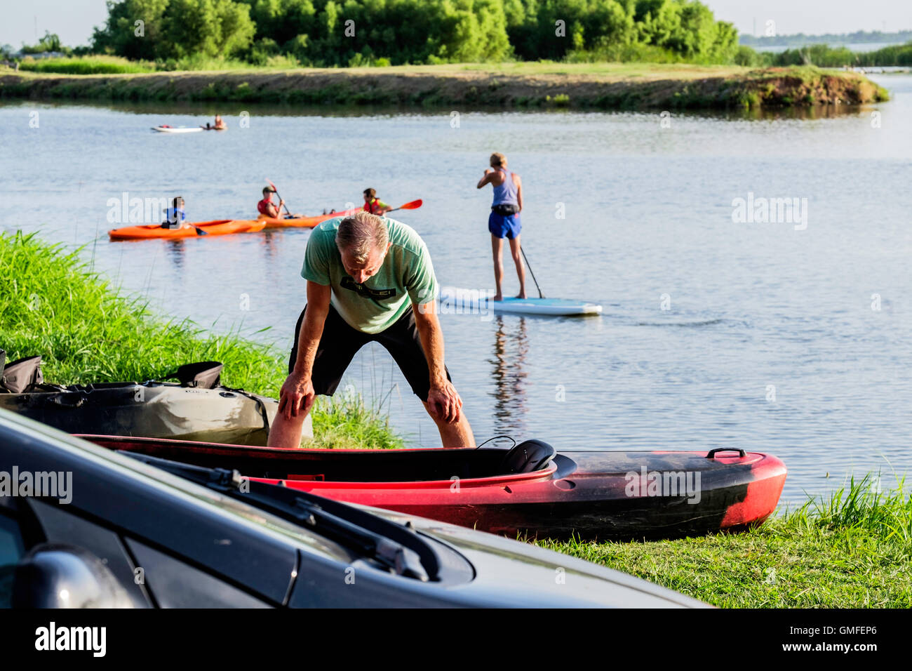 A middle aged Caucasian man looks over his kayak on shore while people play on a paddleboard and kayaks on a river. Stock Photo