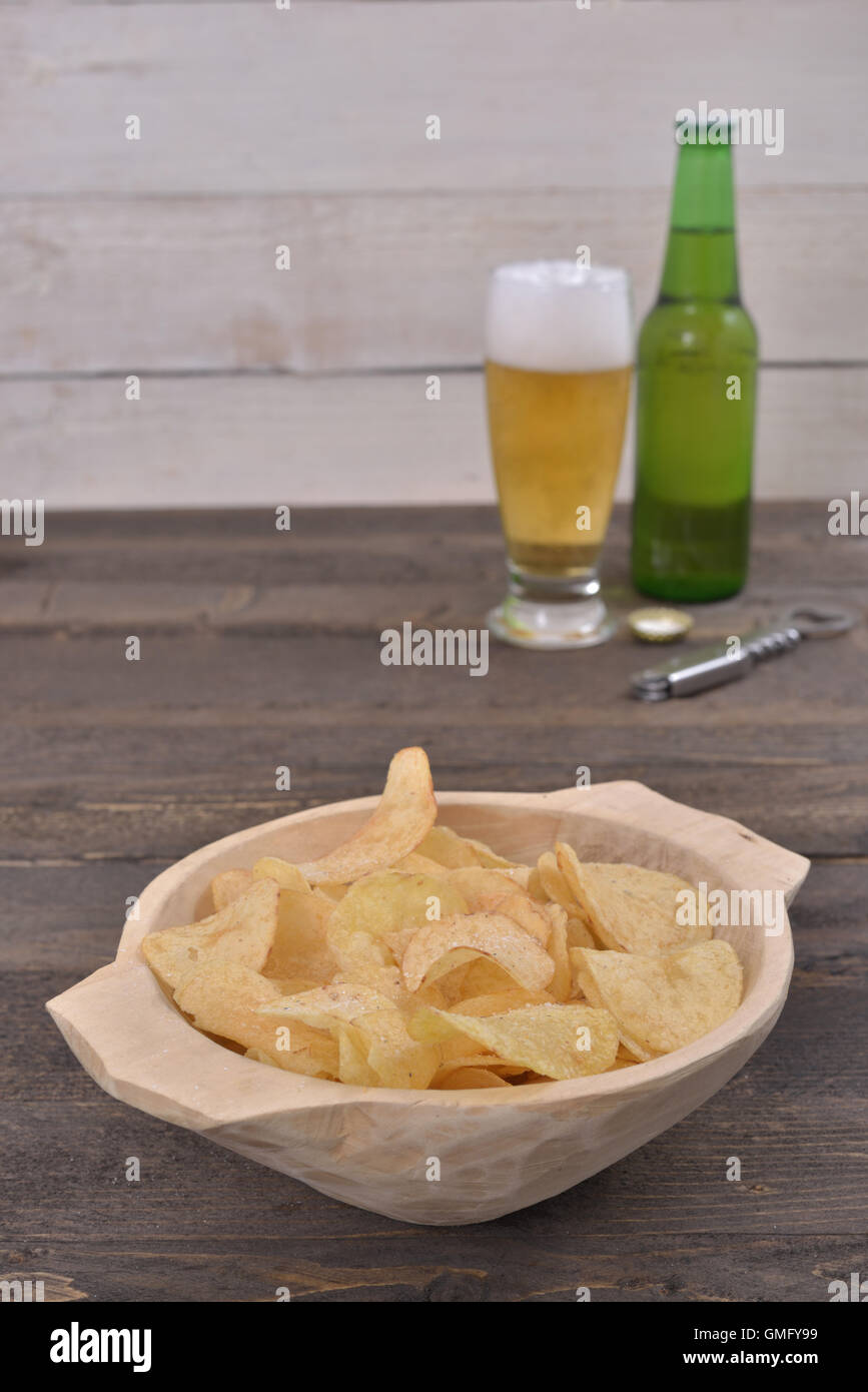 Gold salted potato chips in a wooden bowl on a rustic brown table with a beer glass and a bottle in the background Stock Photo