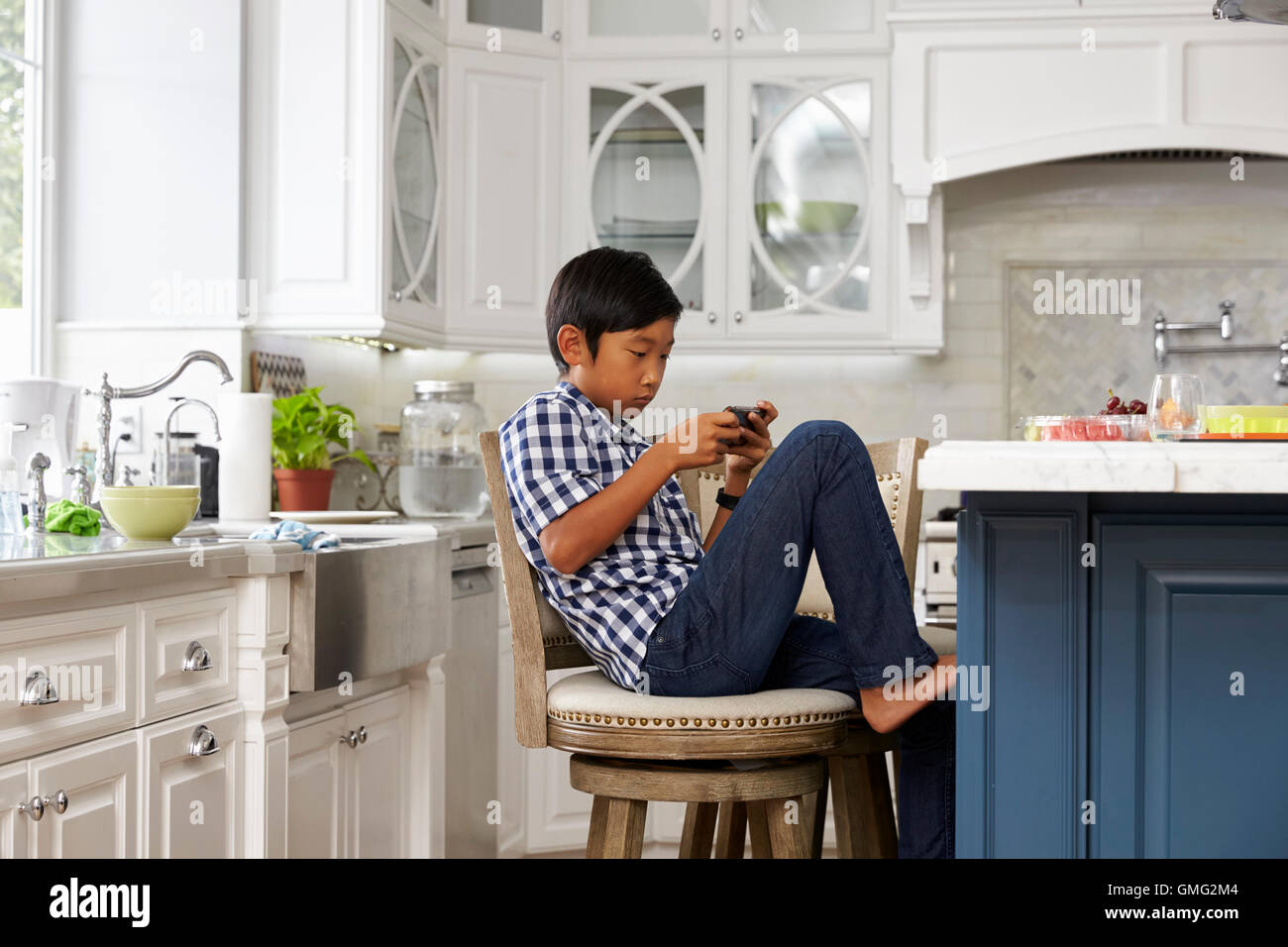 Young Asian Boy Playing Game On Mobile Device In Kitchen Stock Photo