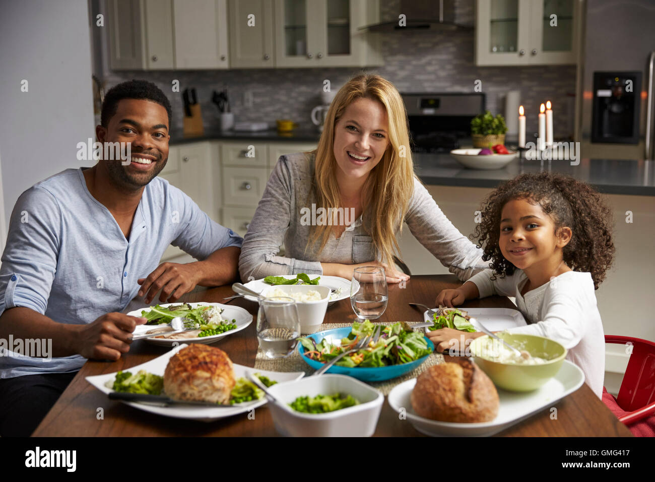 Girl and mixed race parents dine in their kitchen, to camera Stock Photo