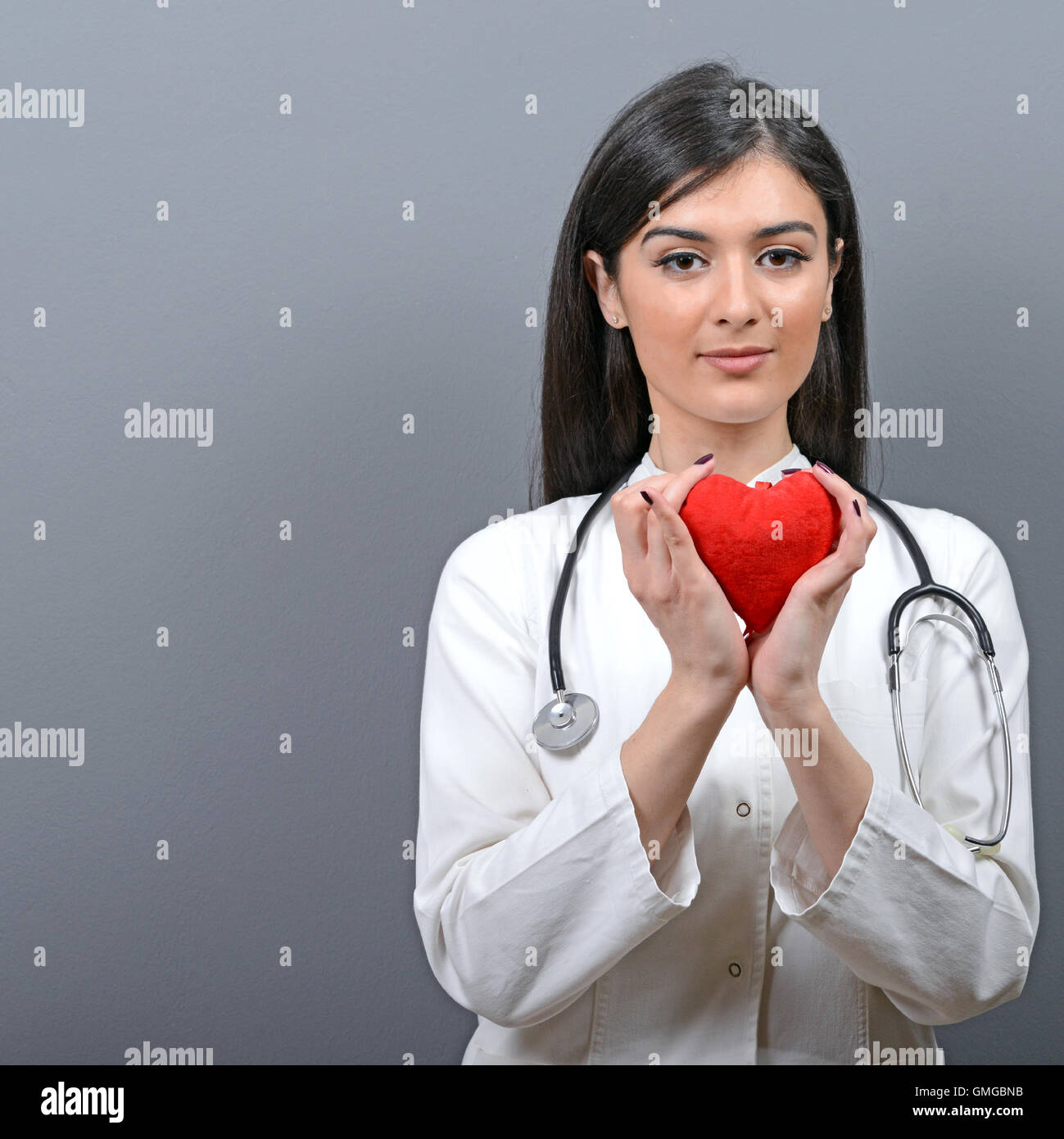 Portrait of young beautiful woman doctor holding red heart against gray ...