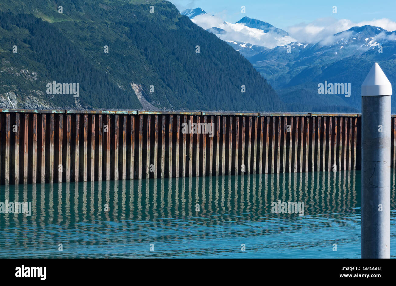 A rusted crenelated sea wall separates its reflection and a forested hillside in Whittier Alaska. Stock Photo