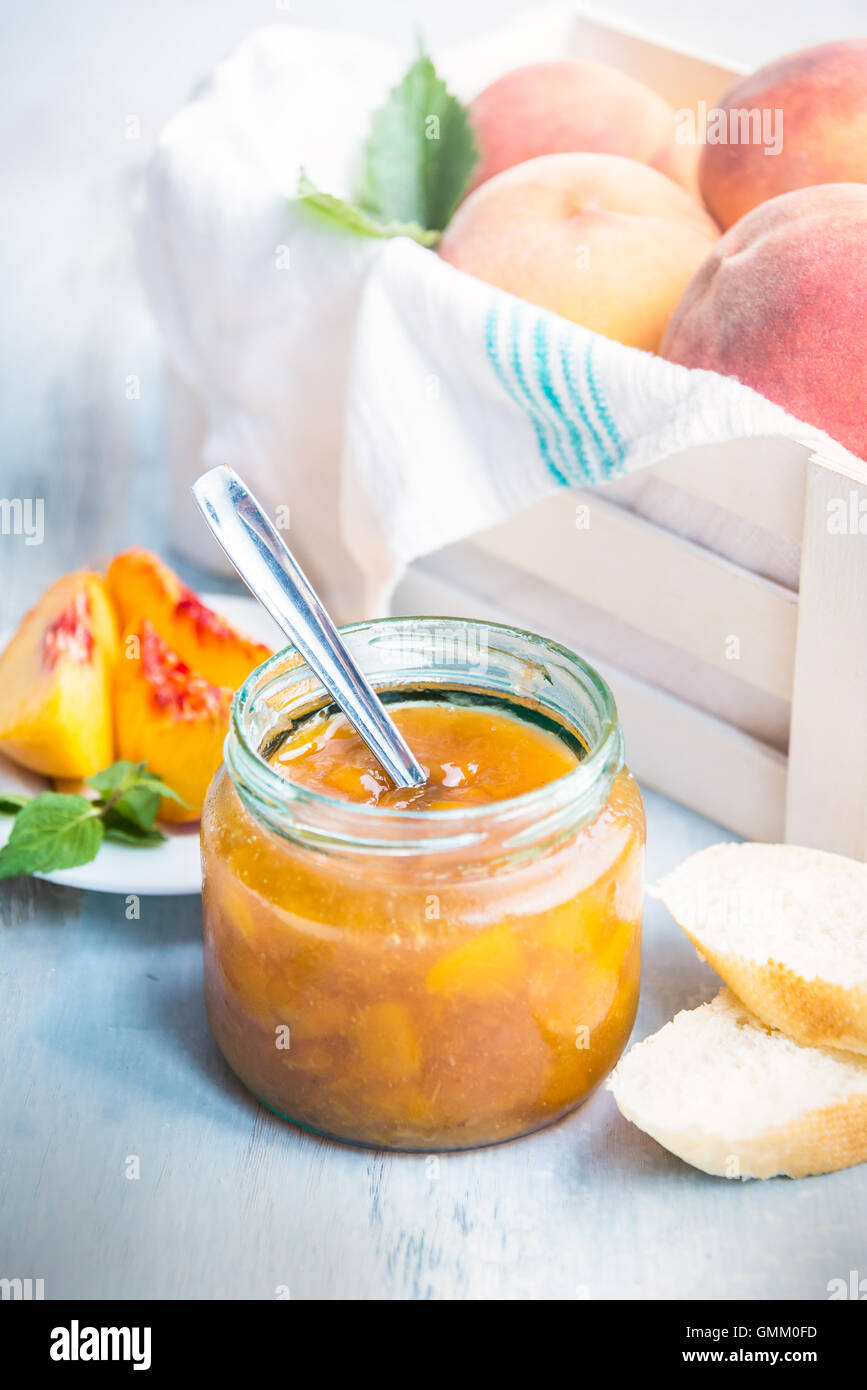 A jar with homemade peach jam in front of a white wood crate full of ripe peaches over a light blue old wood background. Stock Photo