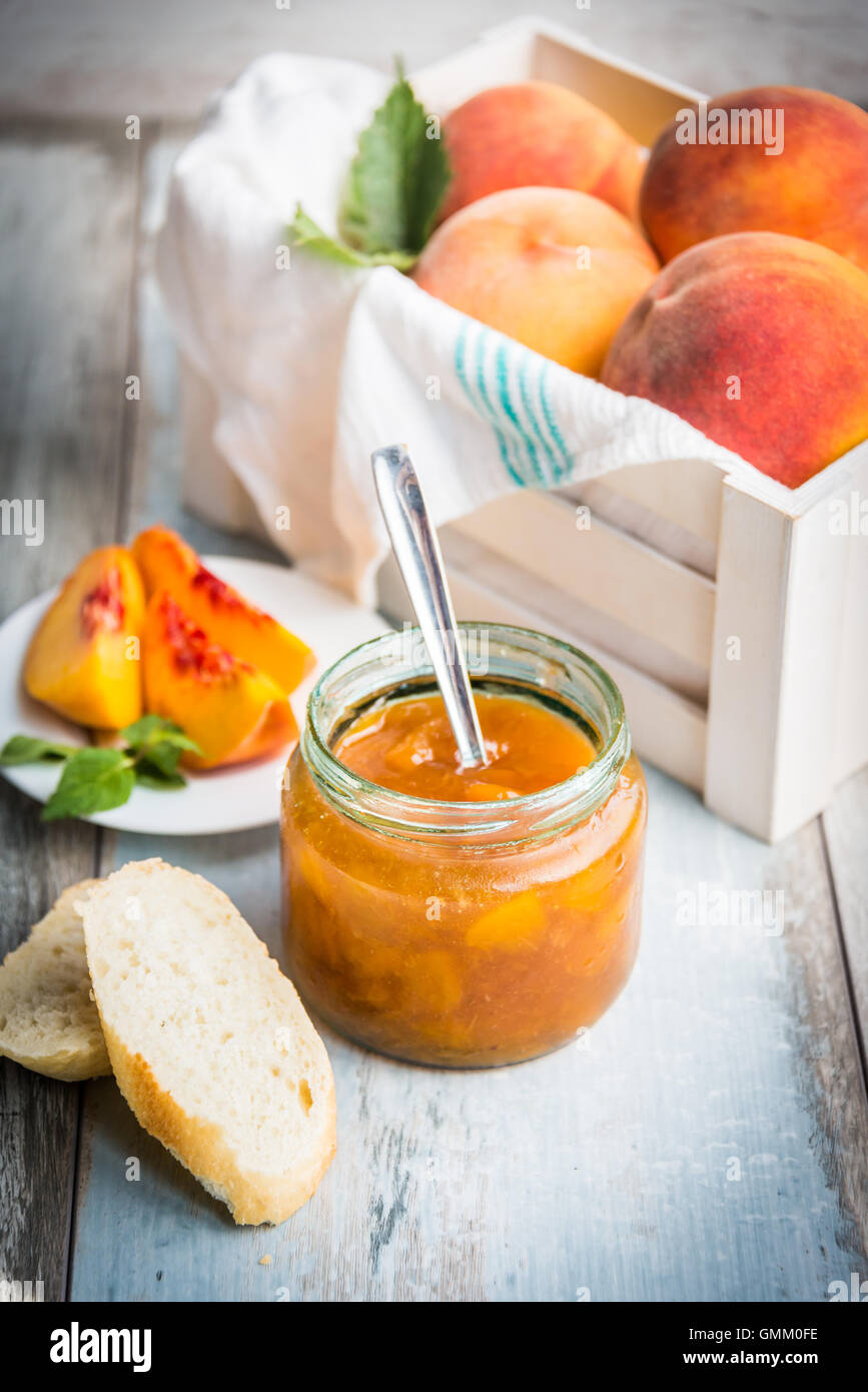 A jar with homemade peach jam in front of a white wood crate full of ripe peaches over a light blue old wood background. Stock Photo