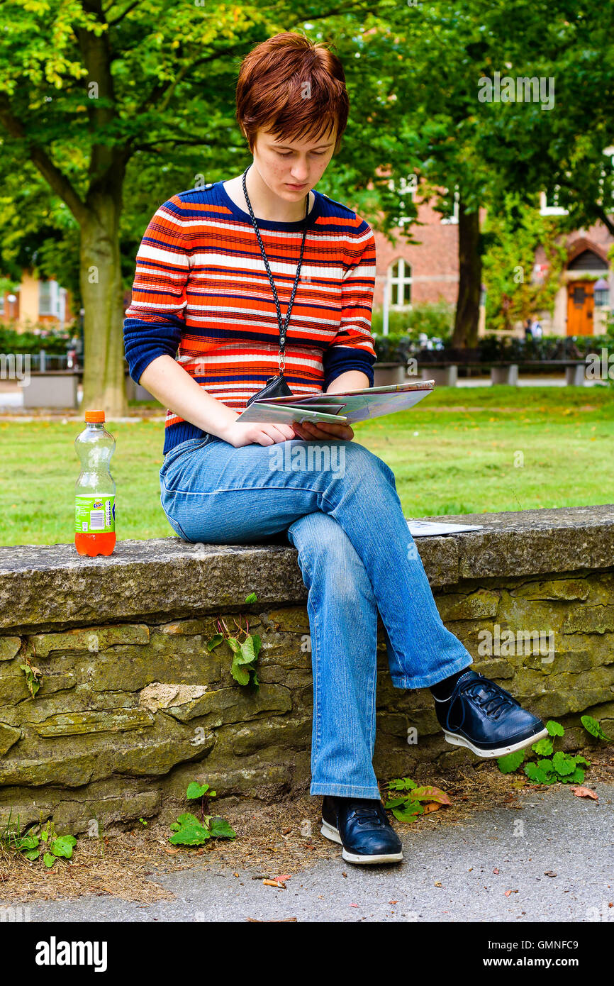 Lund, Sweden - August 24, 2016: Young attractive female sitting on stone wall while looking at a city map. Bottle beside her and Stock Photo