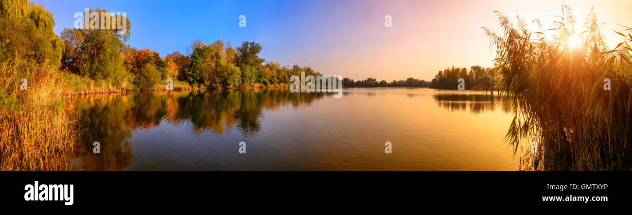 Panorama of a gorgeous sunset at a lake, with gold and blue color and trees reflected in the water Stock Photo