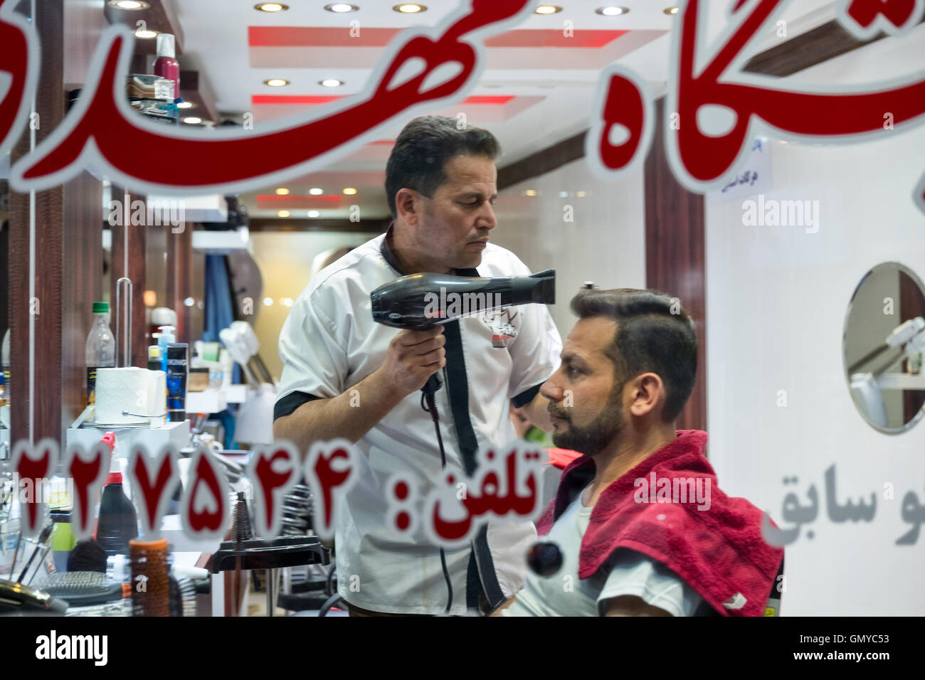 Iranian barber styles man's hair with blow dryer, seen through a window