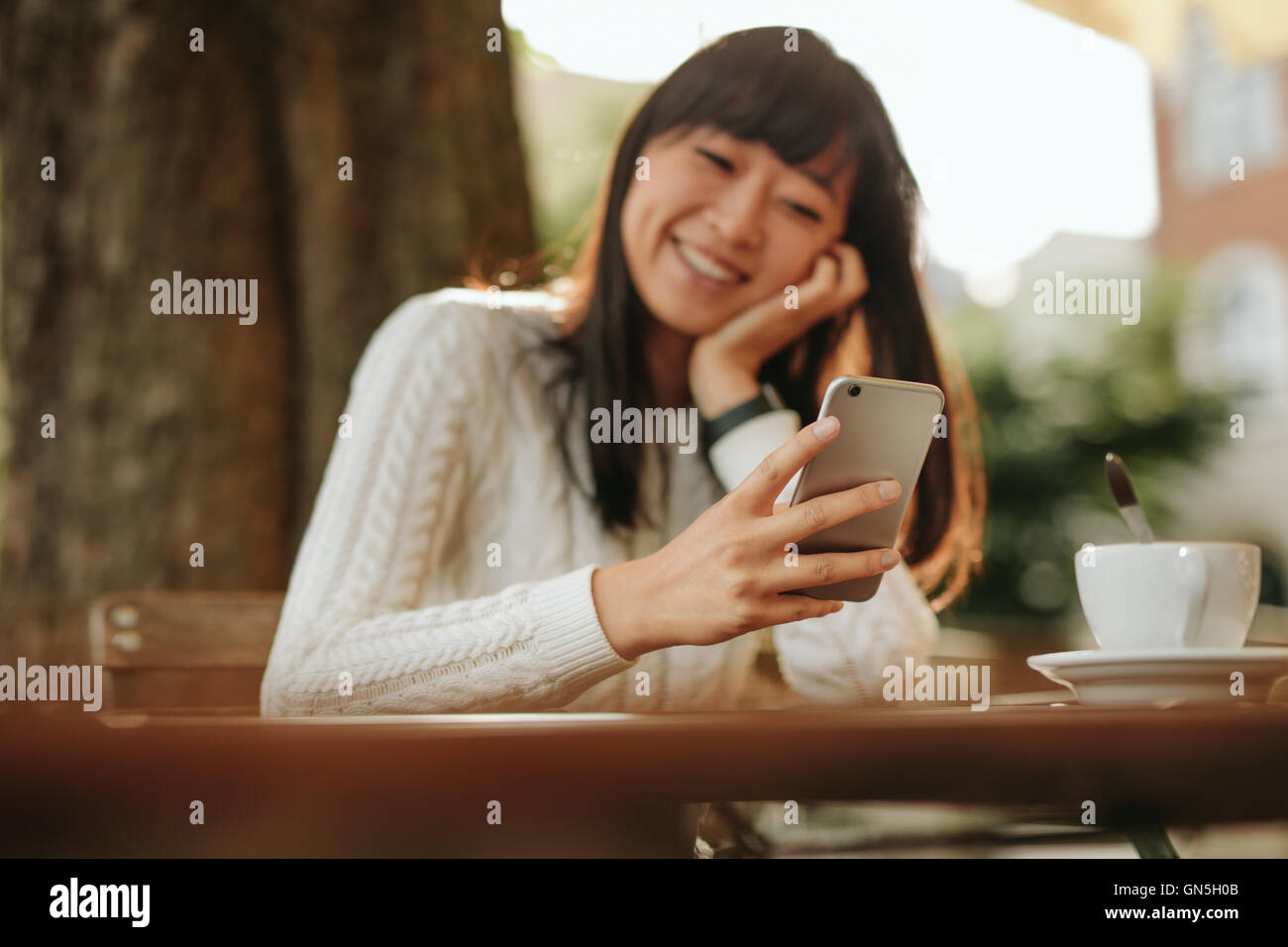 Smiling young woman looking at mobile phone in her hand while sitting at outdoor cafe. Female model reading text message on smar Stock Photo