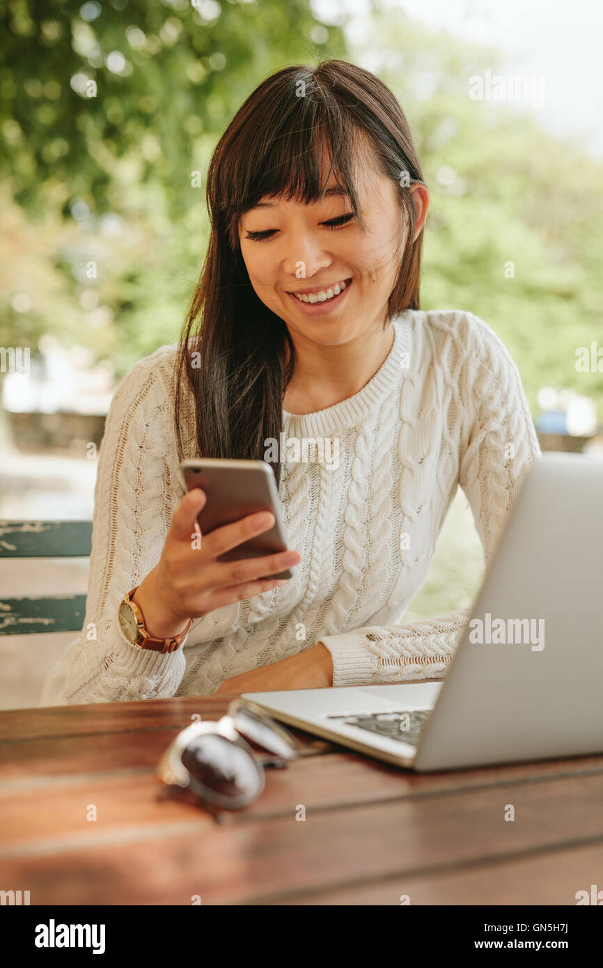 Shot of smiling asian woman using cellphone at outdoor coffee shop. Female sitting at cafe table outdoors using smartphone. Stock Photo