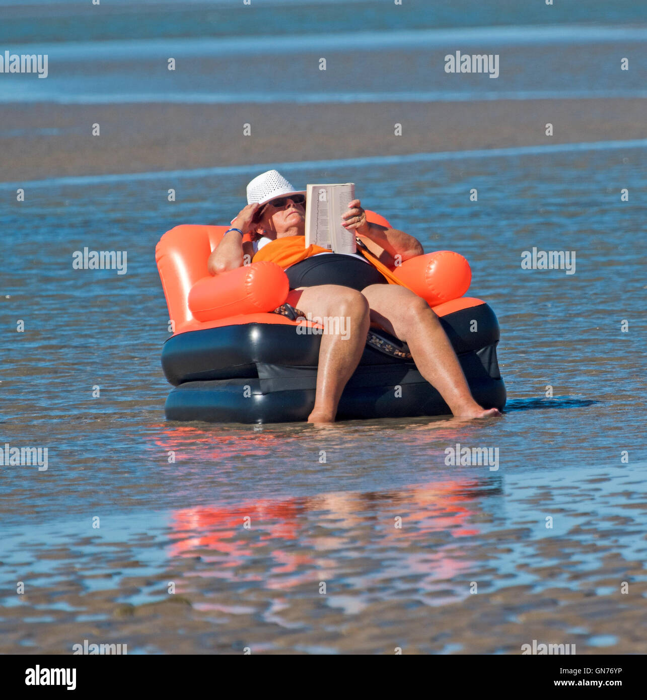 Overweight woman wearing shorts, colourful top & hat seated on inflatable chair reading book at beach in Australia Stock Photo