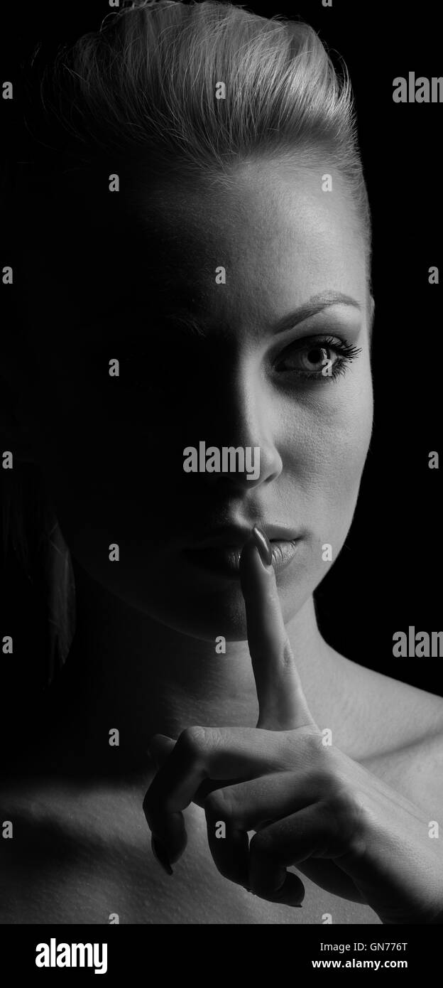 Mystery woman in shadow with finger on lips gesturing silence. Dark portrait. Stock Photo