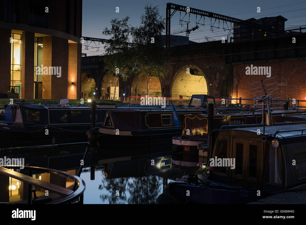 Houseboats moored with the arches under Leeds Railway station and buildings lit up under the late evening sky. Stock Photo