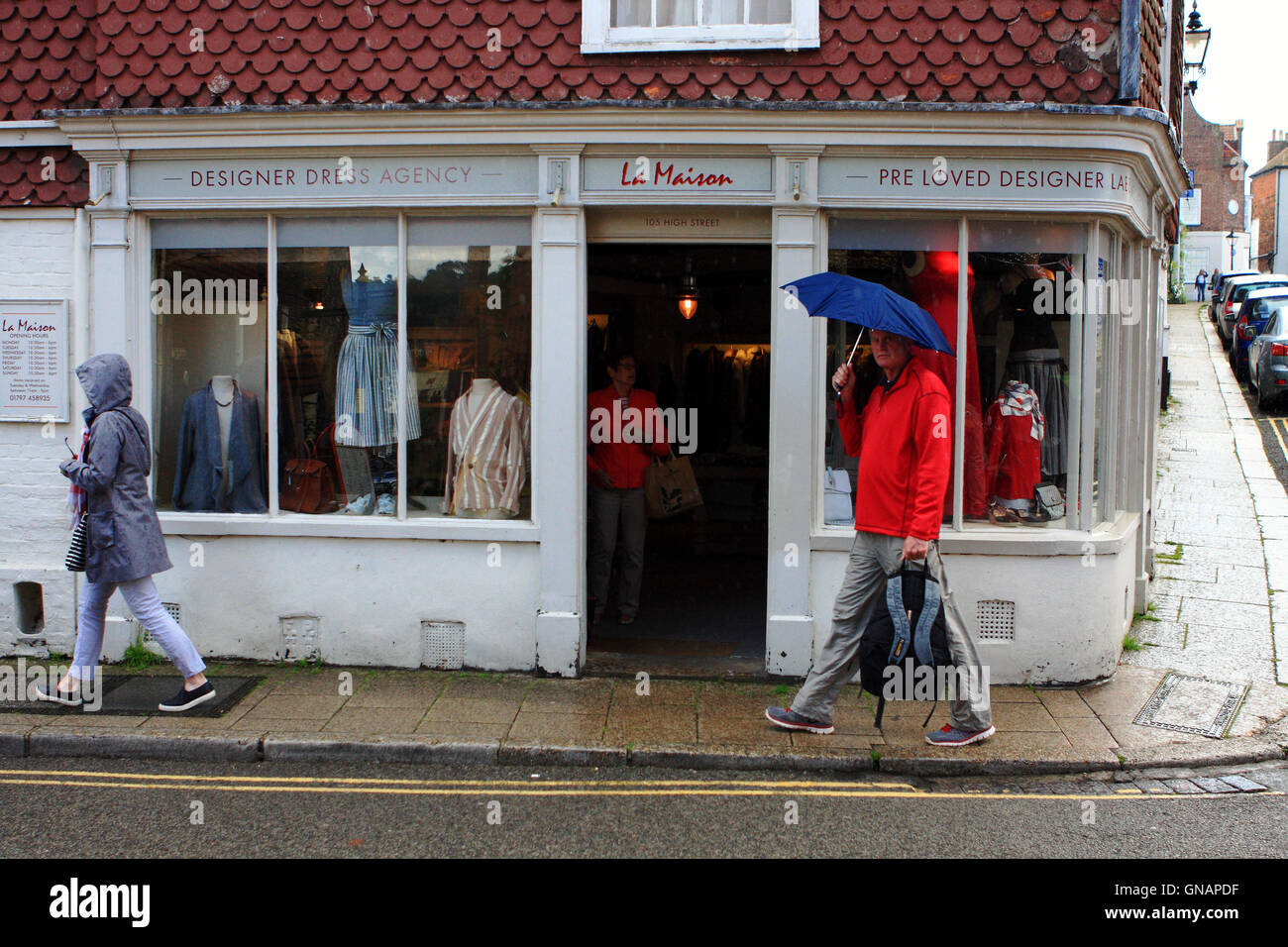 A clothes shop in Rye, East Sussex, England with people walking passed in the rain and a woman approaching the door from inside. Stock Photo