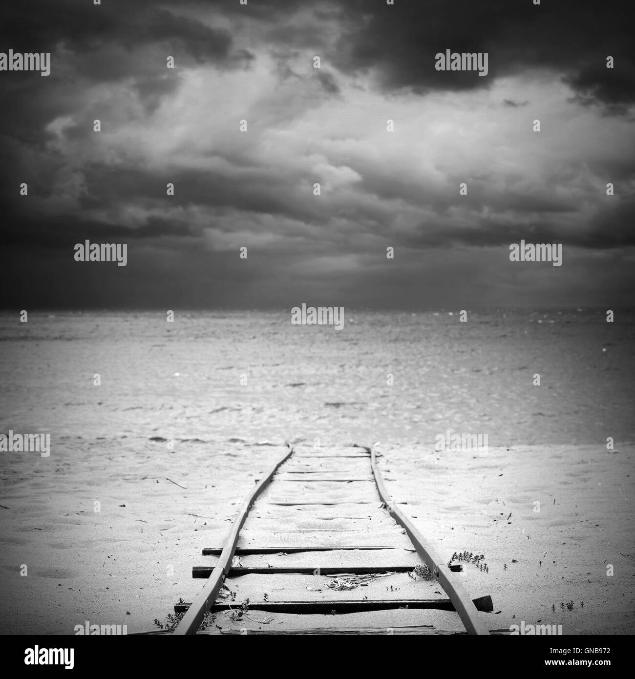 Old railway goes over sandy beach to sea water under dark dramatic cloudy sky, useful for boat launching. Square black and white Stock Photo