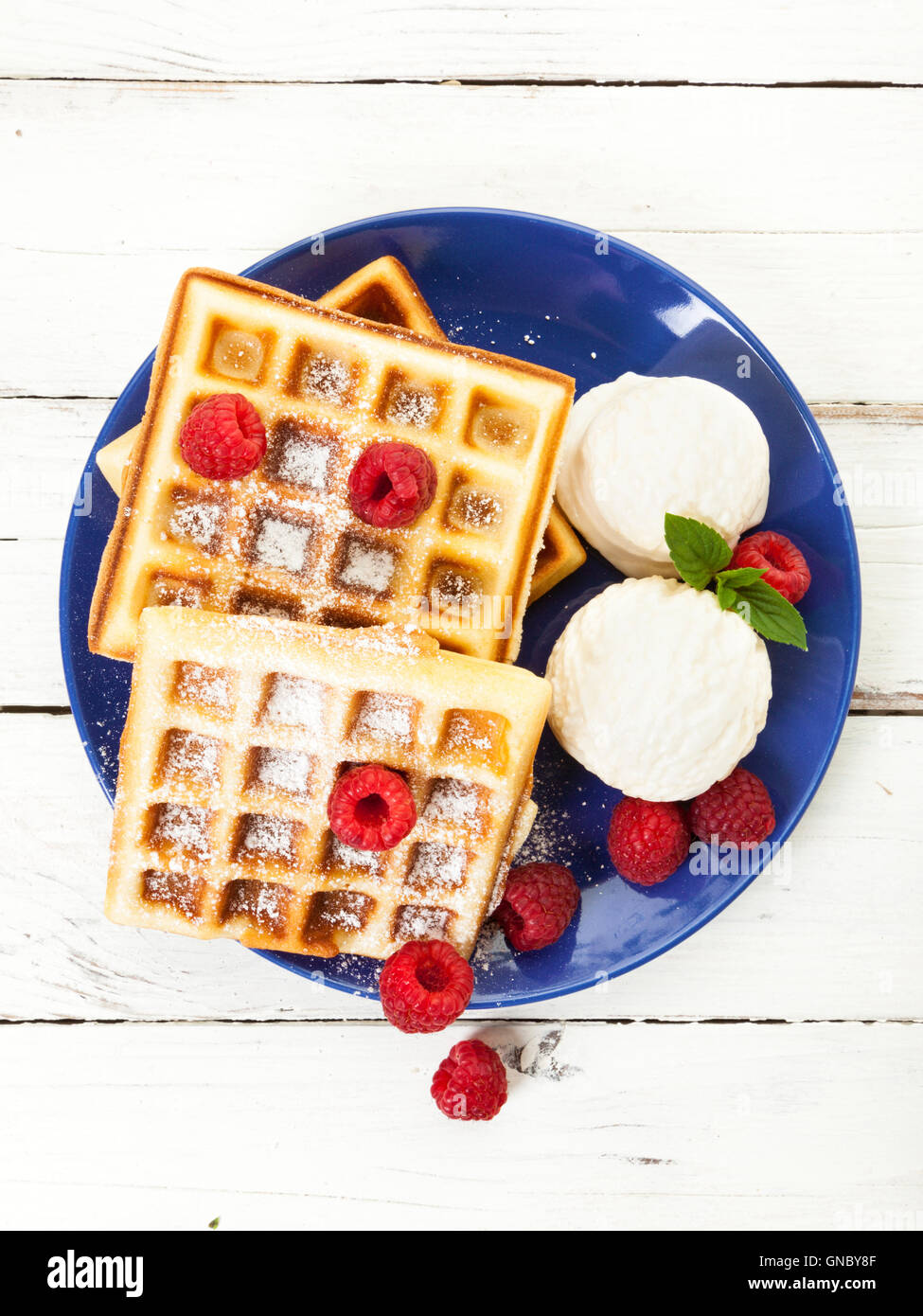 Belgian waffles with raspberries, vanilla ice cream scoops and powdered sugar on blue plate, on rustic wooden table, top view Stock Photo