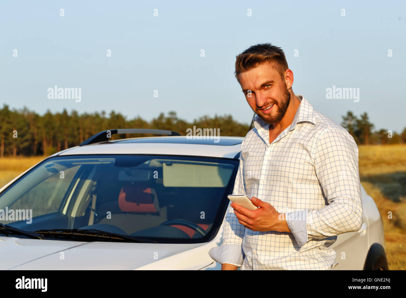 A young handsome man standing in front of the car. A man holding a smartphone and smiles. Always connected. Stock Photo