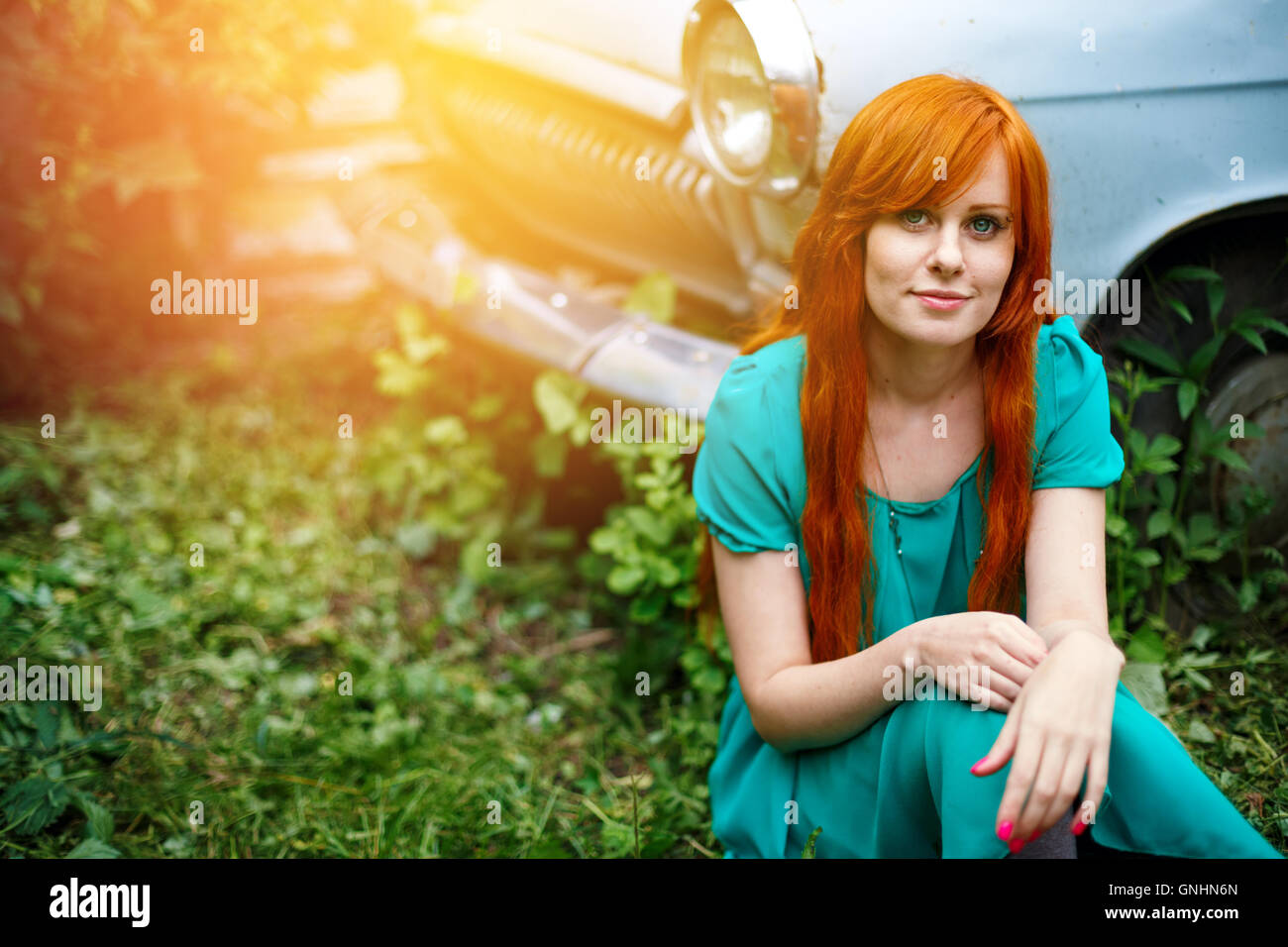 Funny bright young woman posing near vintage abandoned old car. Smiling and sitting in grass. Colored red long hair. Lens flare Stock Photo