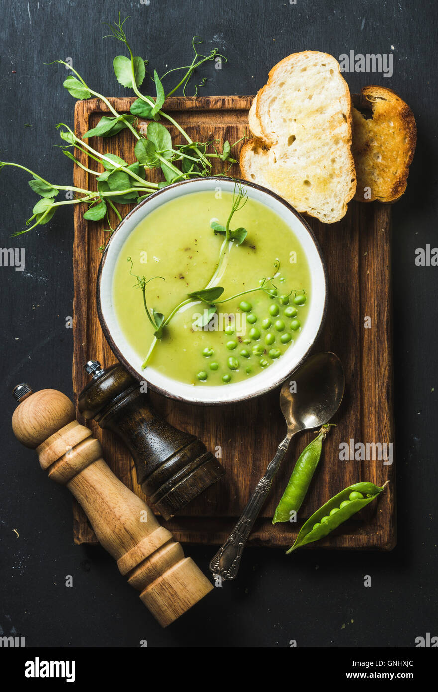 Fresh homemade pea cream soup in white bowl with grilled bread on wooden board over black slate stone backdrop, top view, vertic Stock Photo