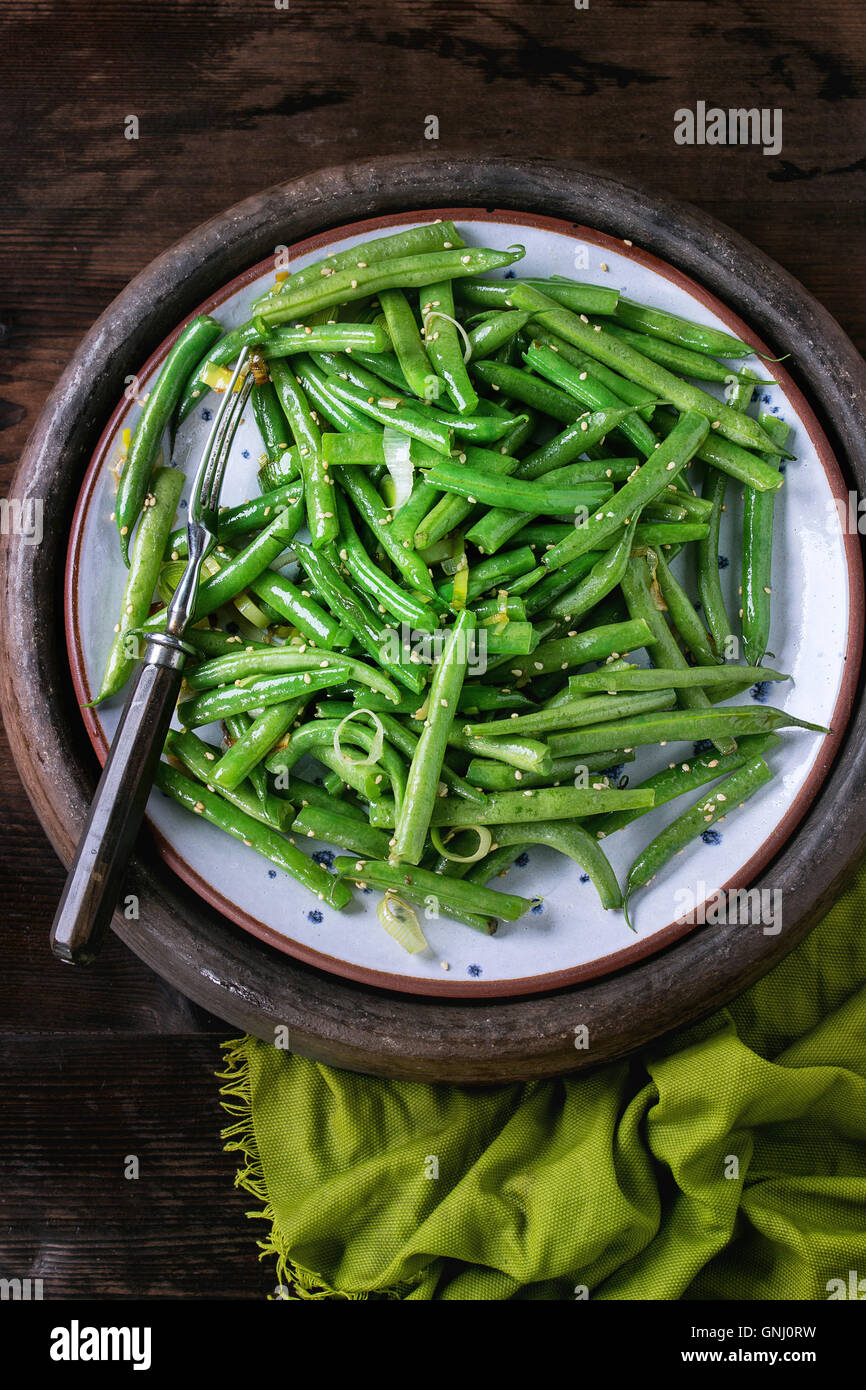 Plate of fried green beans with onion and sesame seeds, served with fork in old clay tray with green textile over dark wooden te Stock Photo