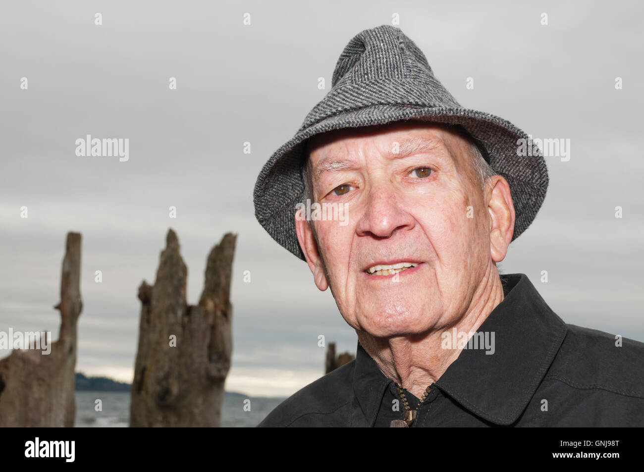 Portrait Senior Man in Gray Tweed Hat and Black Jacket. Background  Moody Grey Sky and Sea. Old Ruins of Pilings. Copy space. Stock Photo