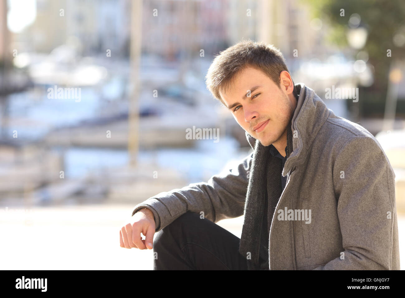 Portrait of a young handsome man wearing a grey jacket in winter sitting in the street of a port Stock Photo