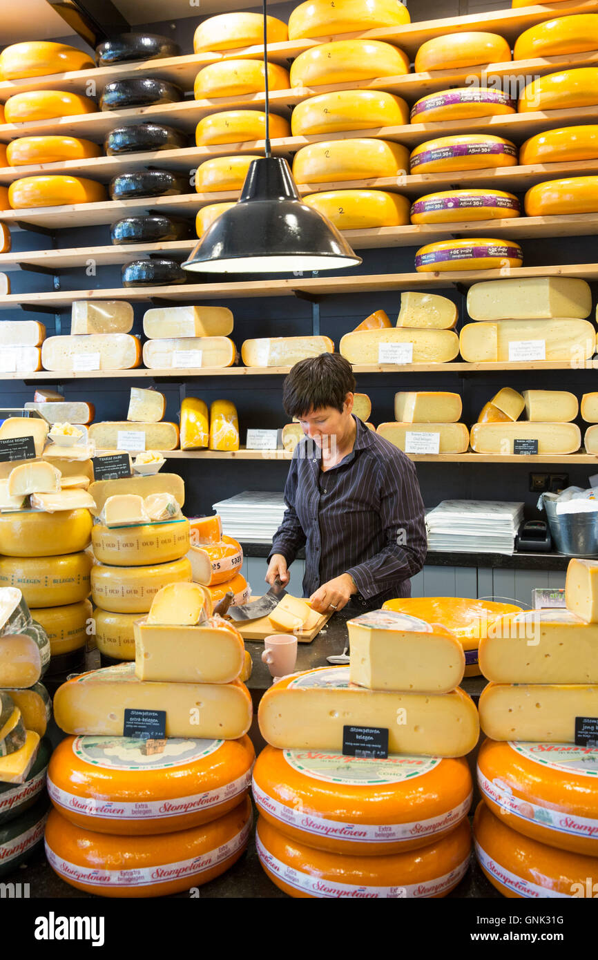 Shelves of cheese wheels and woman cutting cheese wedge at cheese shop 't Kaaswinkeltje in Gouda, Holland, The Netherlands Stock Photo