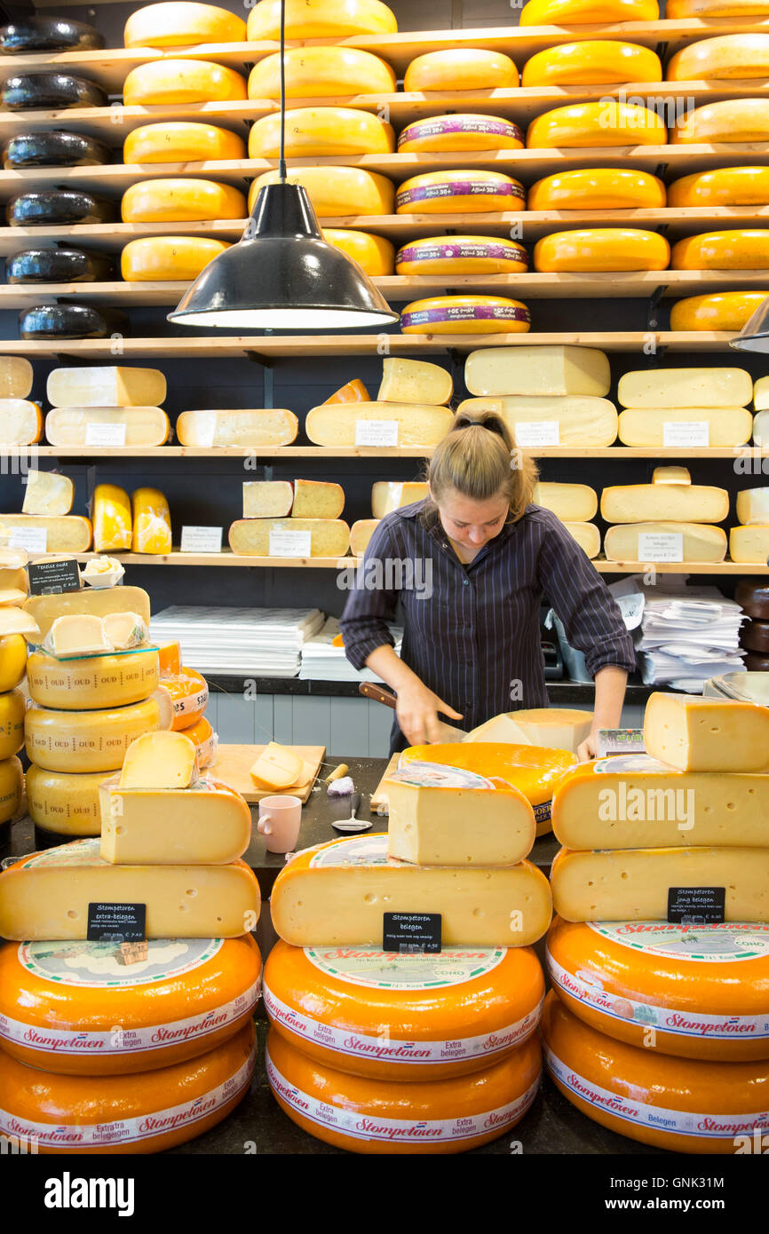 Shelves of cheese wheels and woman cutting cheese wedge at cheese shop 't Kaaswinkeltje in Gouda, Holland, The Netherlands Stock Photo
