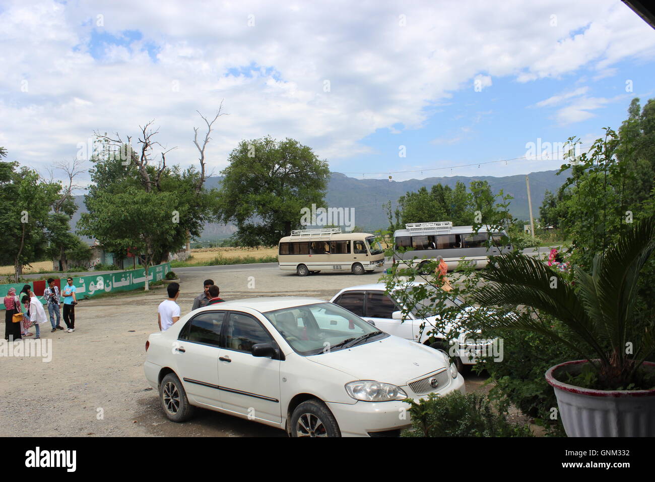 A wide shot of a white car, with two buses in the background, people in groups chatting, and a mountain range in the background. Stock Photo