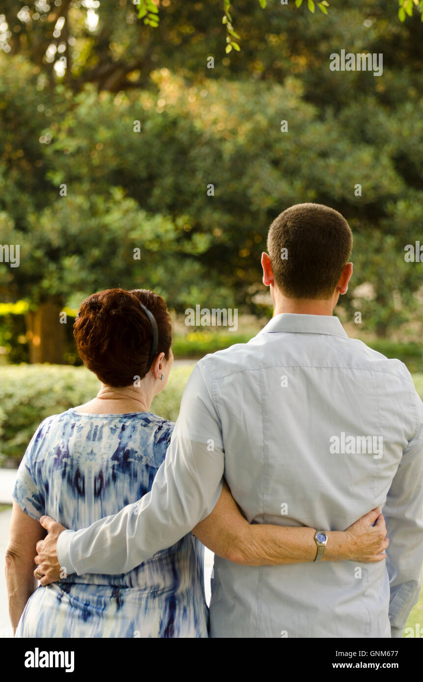 Mother and adult son walking in a park with their arms around each other as the sun is setting casting a golden hue on them. Stock Photo