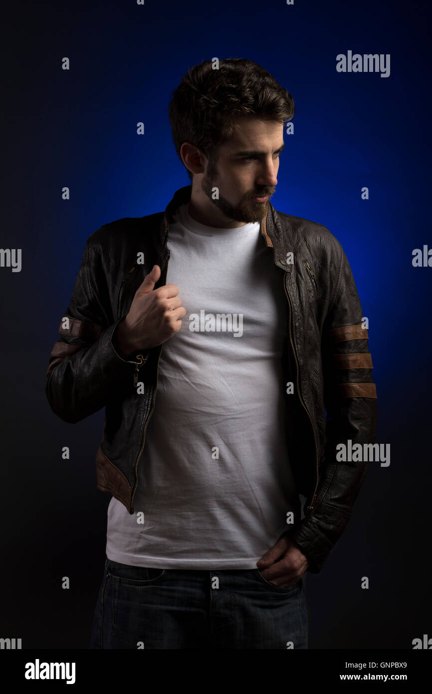 Male model with groomed beard wearing white tee and leather jacket with blue backdrop in studio Stock Photo